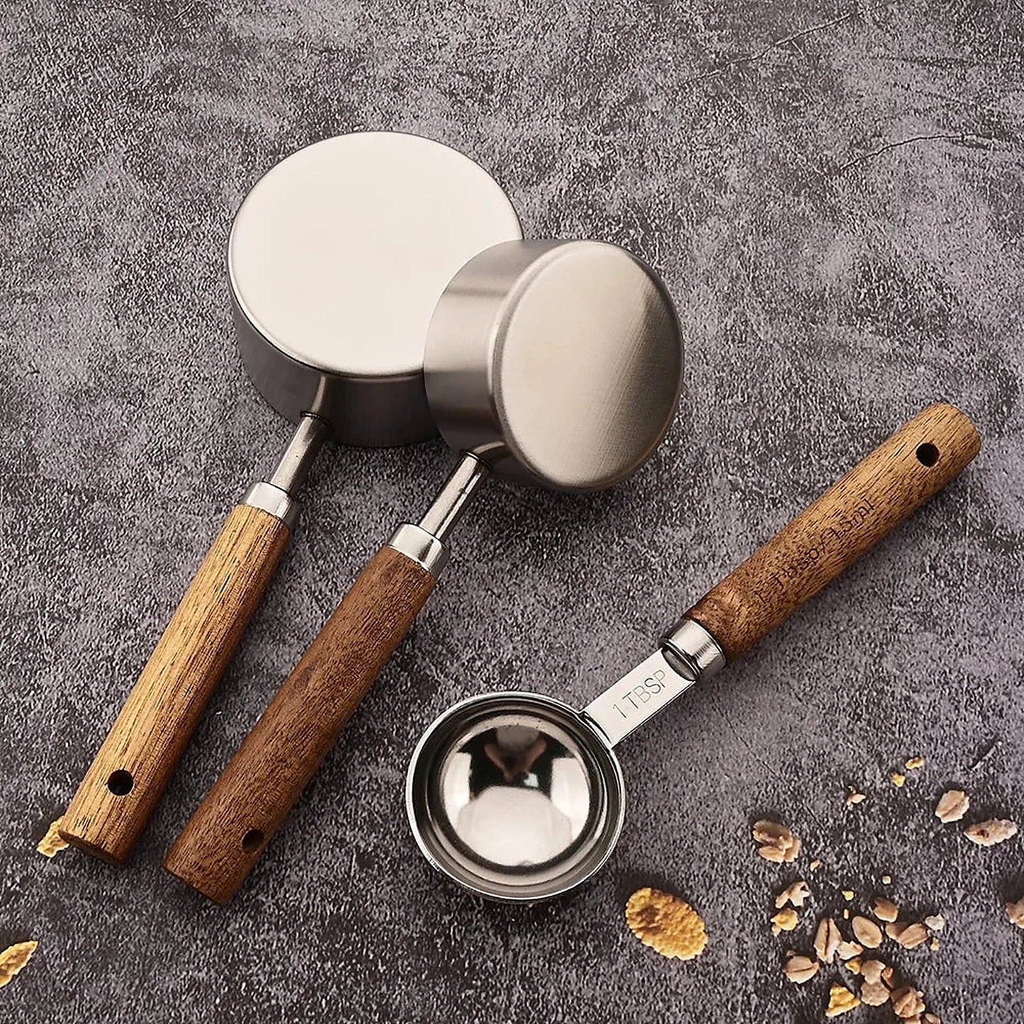 Tasse à mesurer en inox à manche en bois et tasse doseuse, sur une surface grise avec des grains de café.