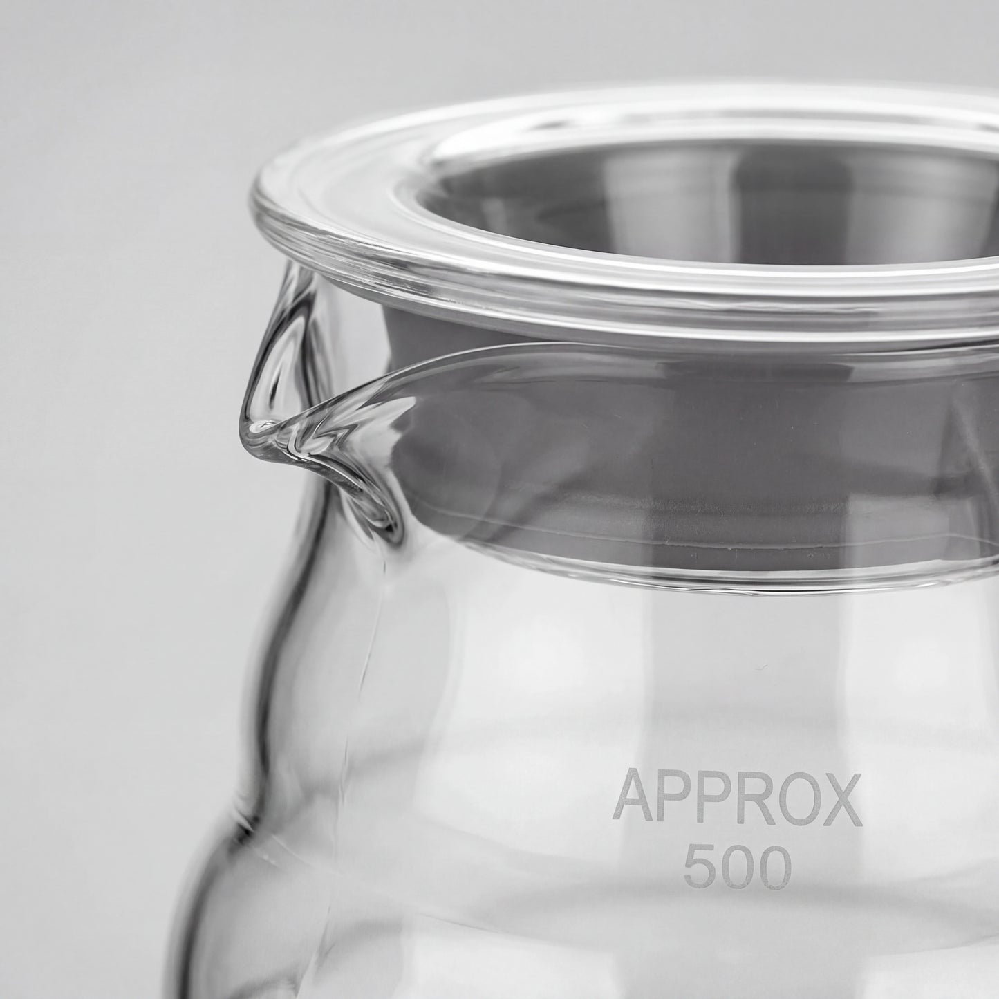 Gros plan du bec verseur et du couvercle en verre à liseré inox d'une carafe à café.