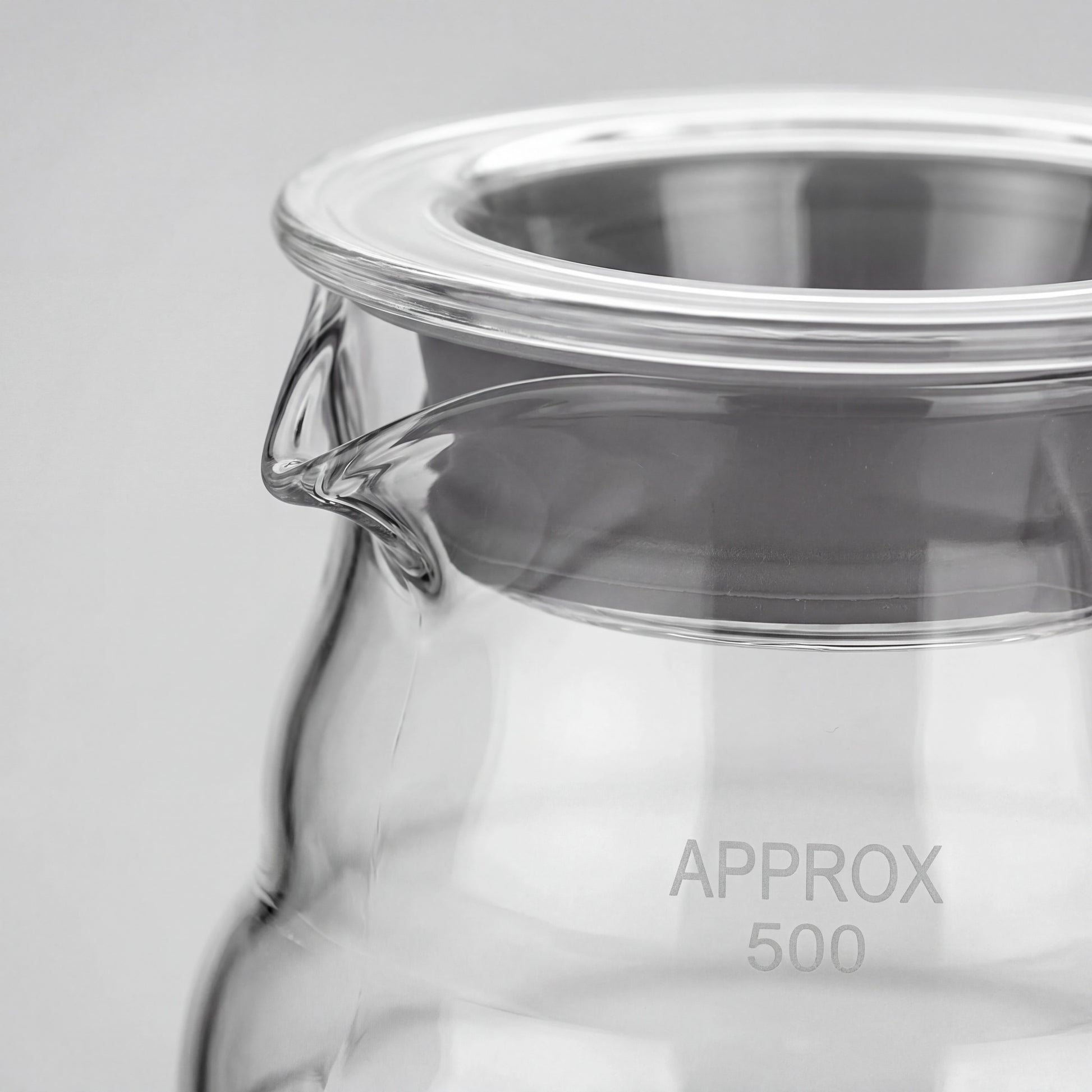 Gros plan du bec verseur et du couvercle en verre à liseré inox d'une carafe à café.