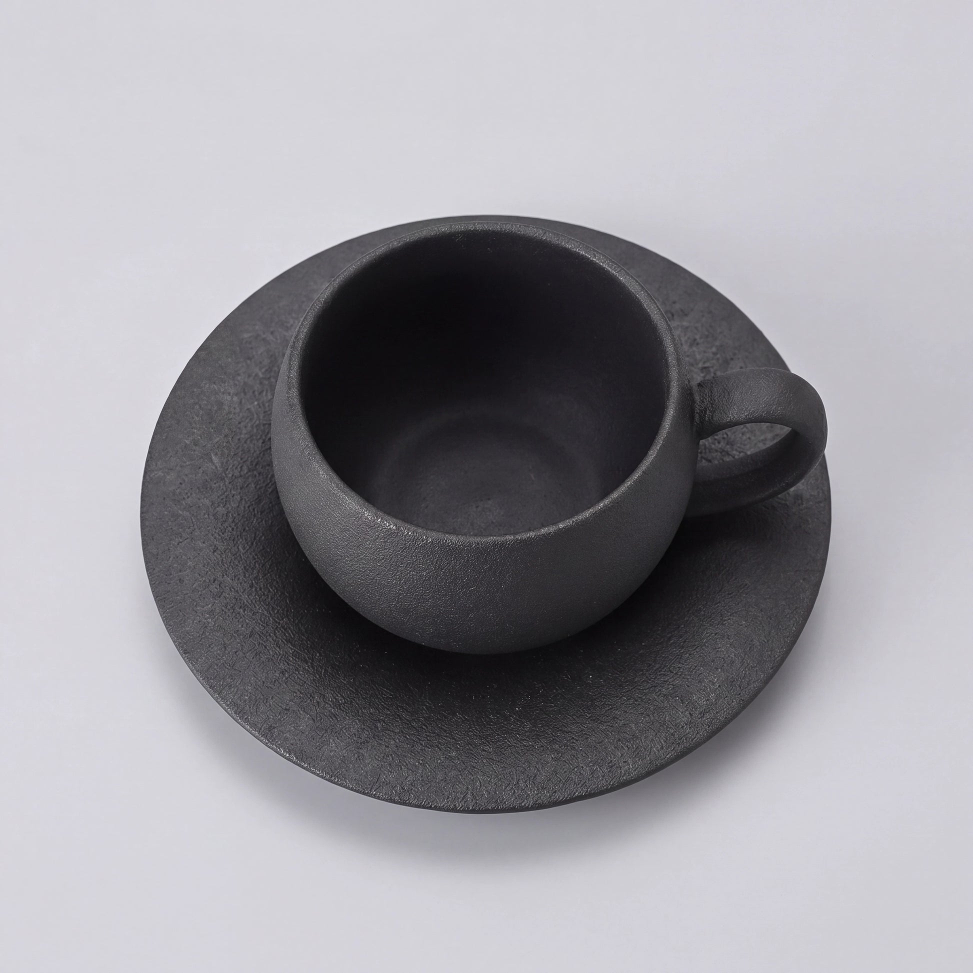 Tasse à café en céramique noire et sa soucoupe vues en plongée, intérieur visible.