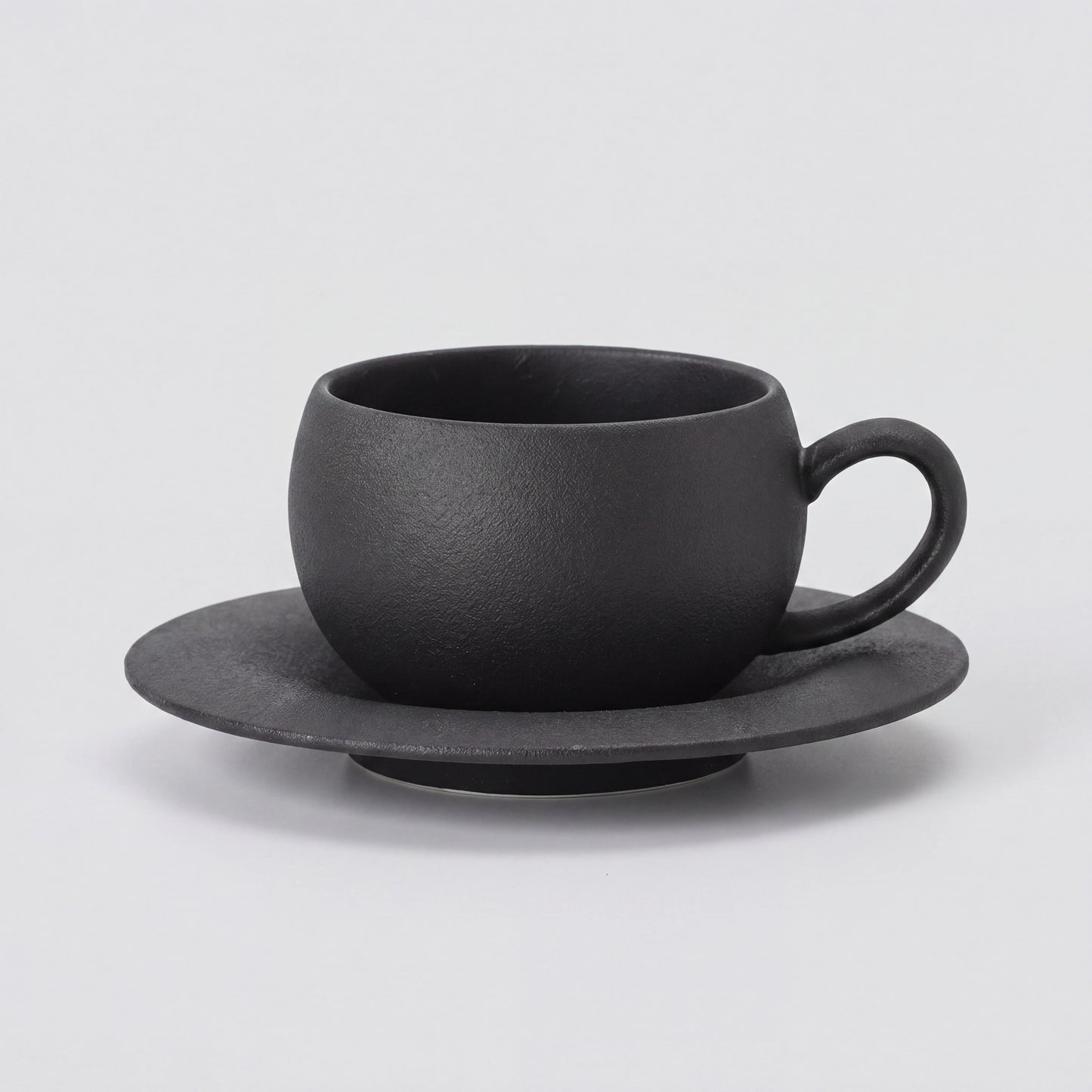 Tasse à café en céramique noire texturée et sa soucoupe, vue de profil sur fond clair.
