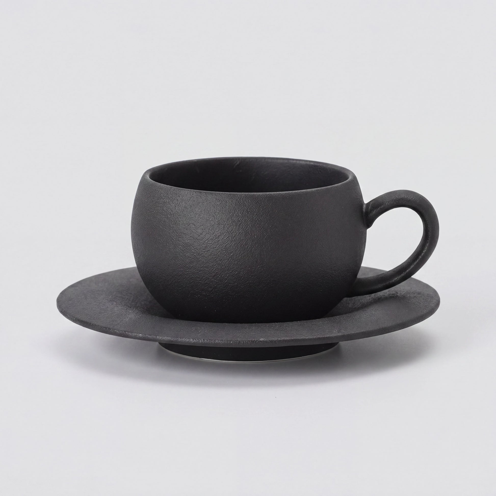 Tasse à café en céramique noire texturée et sa soucoupe, vue de profil sur fond clair.