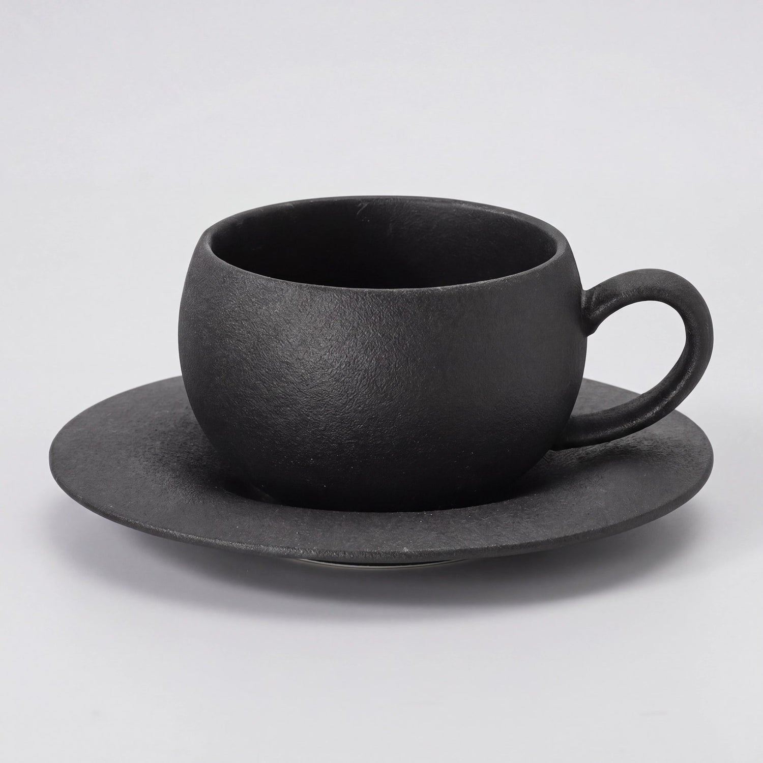 Tasse à café en céramique noire texturée avec sa soucoupe, vue de trois-quarts sur fond clair.