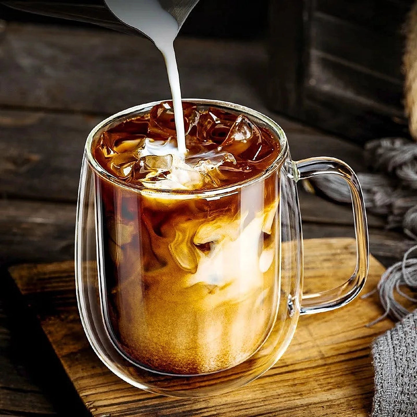 Verre à café double paroi avec poignée contenant un café glacé, lait versé, sur une planche en bois.